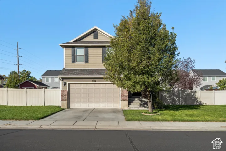 Two-story exterior featuring tan horizontal siding, a two-car garage, brick facade accents, and a fenced front yard with mature landscaping