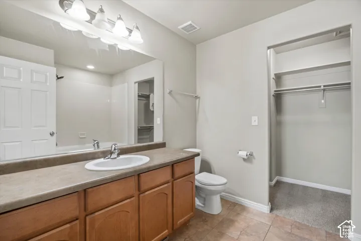 Bathroom featuring a wood-finish vanity with a stone-surface countertop, an oval undermount sink, a full-width vanity mirror, tile flooring, and a walk-in closet with built-in shelving