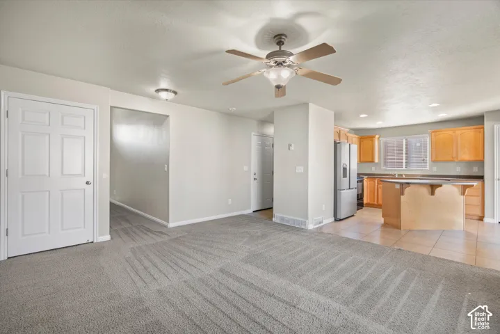 Interior space featuring neutral carpeting, a ceiling fan with integrated lighting, kitchen cabinetry, a kitchen island, and tile flooring