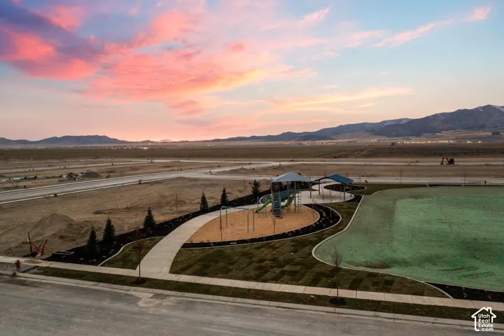 Community park featuring a structured playground, covered pavilion, concrete pathways, manicured lawn areas, and mountain backdrop