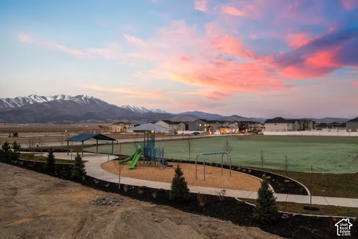 Community park featuring a playground, metal-roofed pavilion, concrete walking paths, and landscaped perimeter with evergreen trees, framed by a mountain backdrop