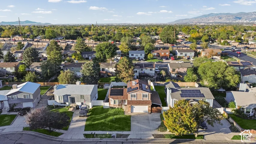 Aerial perspective of a multi-story residence featuring a two-car garage, roof-mounted solar panels, paved driveway, and grass lawn
