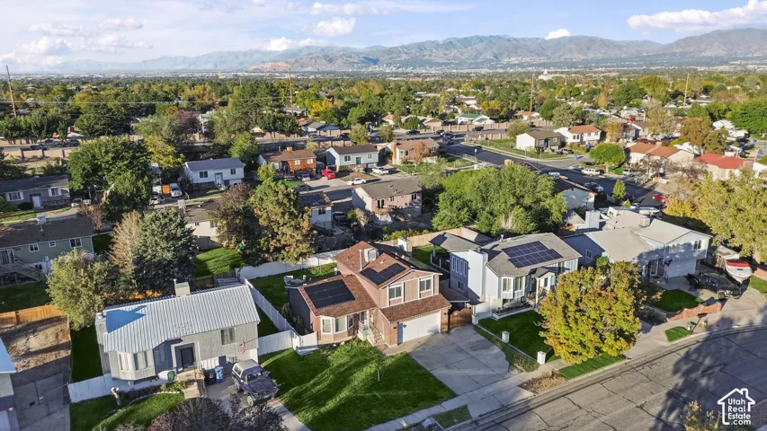 Aerial perspective featuring residential lots with manicured lawns, mature landscaping, roof-mounted solar panels, and a mountain range backdrop