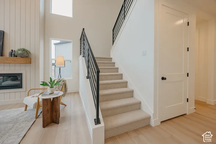 Staircase with carpeted steps and black metal horizontal railing, featuring high ceilings, vertical shiplap wall accent, inset fireplace with wood mantle, and light-toned hard surface flooring