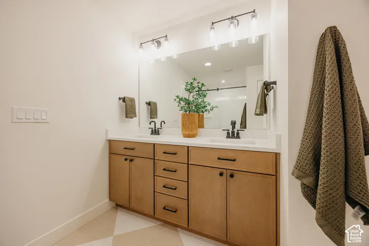 Dual-sink vanity with light-tone wood cabinetry, solid-surface countertops, matte black hardware, and two wall-mounted three-light fixtures