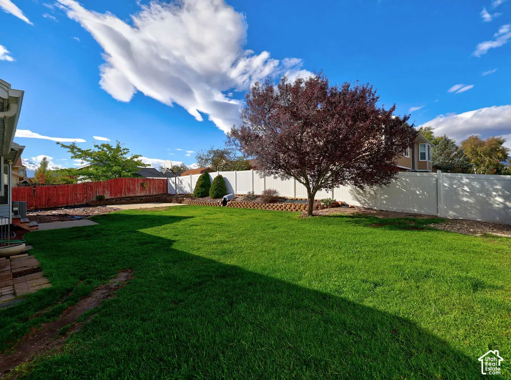 Backyard featuring a manicured lawn, tiered stone retaining walls, mature landscaping, and white vinyl privacy fencing