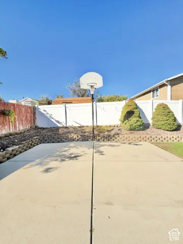 Concrete sports court, white vinyl perimeter fencing, tiered stone retaining walls, and established evergreen shrubbery