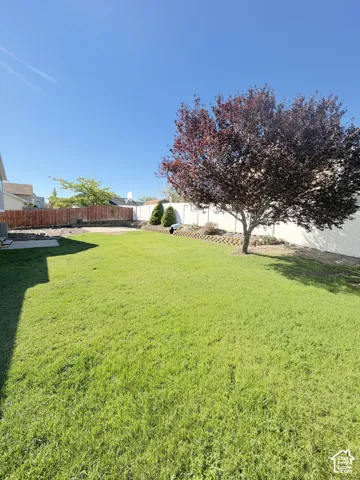 Backyard featuring a manicured lawn, perimeter fencing, mature tree, stone-look retaining wall, and concrete patio