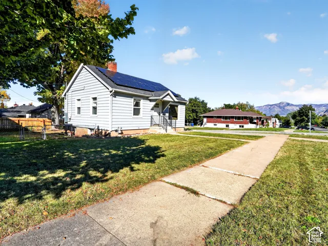 Light-colored horizontal siding exterior featuring roof-mounted solar panels, a brick chimney, and concrete walkway, set against a mountain backdrop