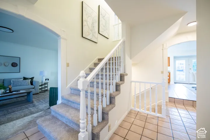 Staircase featuring white balusters and newel posts, complemented by arched architectural entryways, ceramic tile flooring, and carpeted stair treads