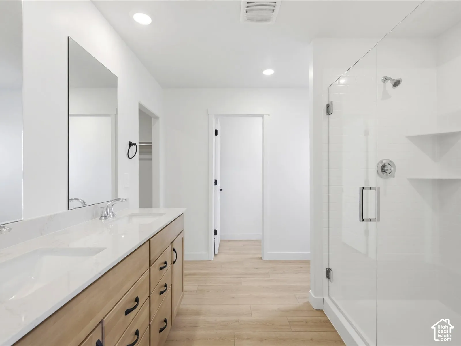 **DISCLAIMER**  Interior package may vary. Double vanity with a light-finish wood cabinet, white stone-surface countertop, frameless glass shower enclosure, and wood-finish flooring