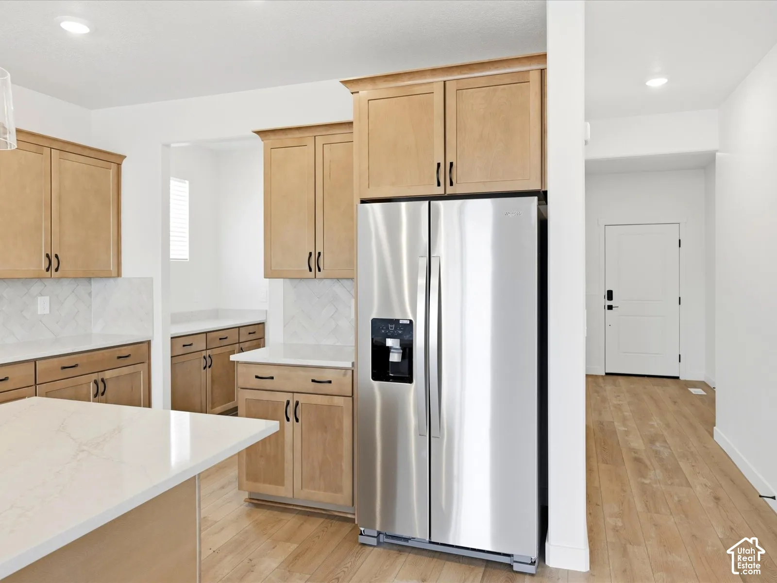 **DISCLAIMER**  Interior package may vary. Kitchen featuring light-tone wood cabinetry, stone countertops, herringbone-pattern tile backsplash, stainless steel refrigerator, and wood-finish flooring