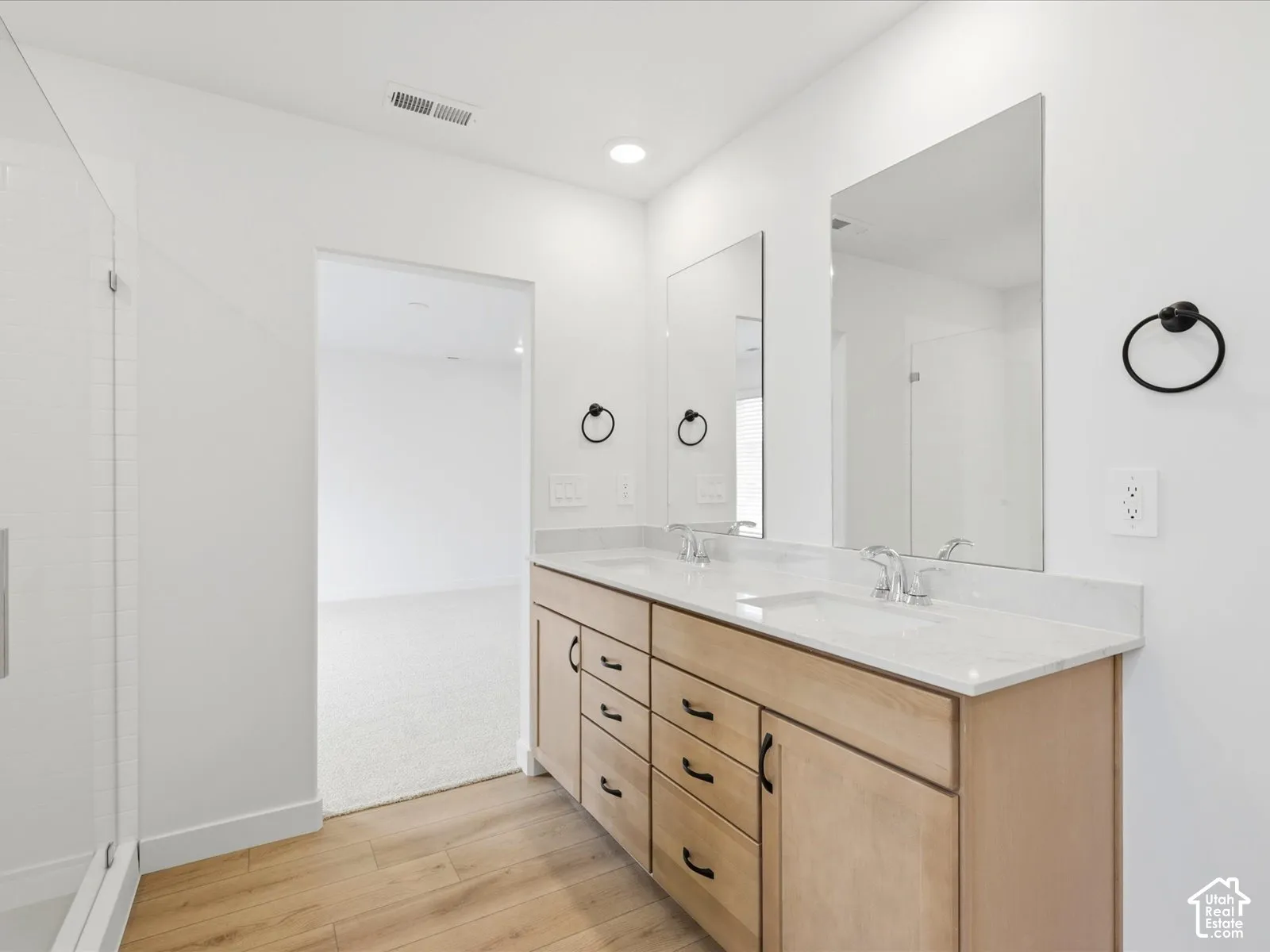 **DISCLAIMER**  Interior package may vary. Dual-sink vanity featuring light wood cabinetry, white stone-finish countertops, matte black hardware, and large frameless vanity mirrors, complemented by glass-enclosed shower with white tiled surround and wood-
