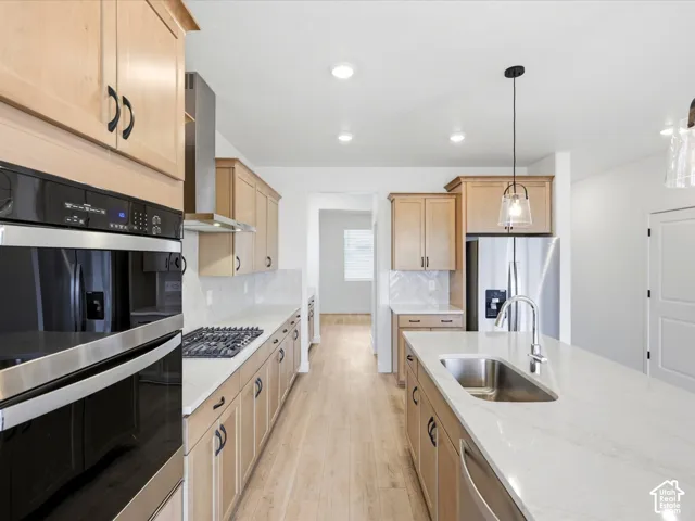 **DISCLAIMER**  Interior package may vary. Kitchen featuring light wood-finish cabinetry, stone-surface countertops, a stainless steel gas cooktop with a chimney-style hood, a built-in double oven, and a center island with an undermount sink