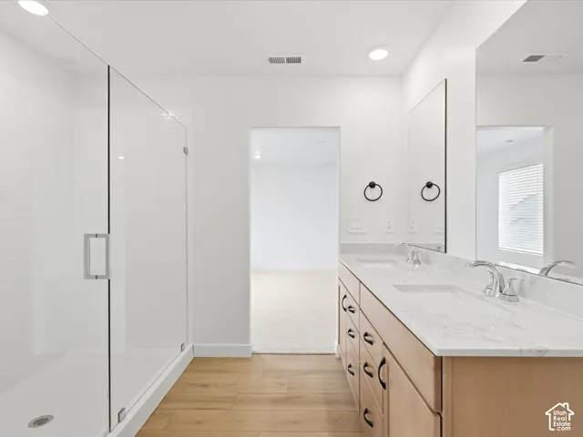 **DISCLAIMER**  Interior package may vary. Bathroom featuring a glass-enclosed shower, wood-finish flooring, a double vanity with stone-style countertops, integrated dual sinks, and matte black hardware