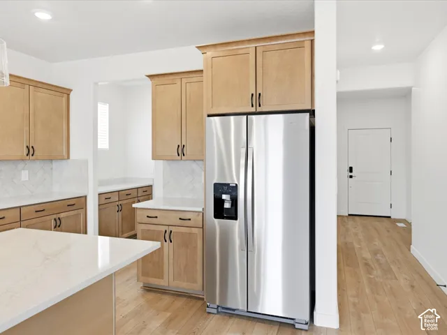 **DISCLAIMER**  Interior package may vary. Kitchen featuring light-tone wood cabinetry, stone countertops, herringbone-pattern tile backsplash, stainless steel refrigerator, and wood-finish flooring