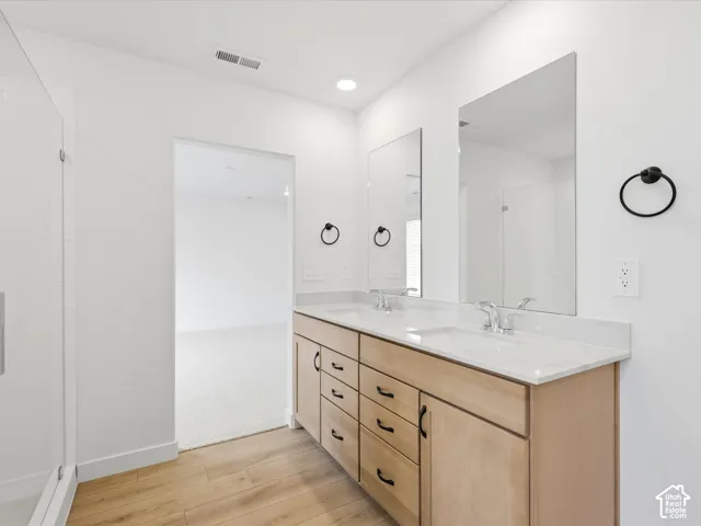 **DISCLAIMER**  Interior package may vary. Dual-sink vanity featuring light wood cabinetry, white stone-finish countertops, matte black hardware, and large frameless vanity mirrors, complemented by glass-enclosed shower with white tiled surround and wood-