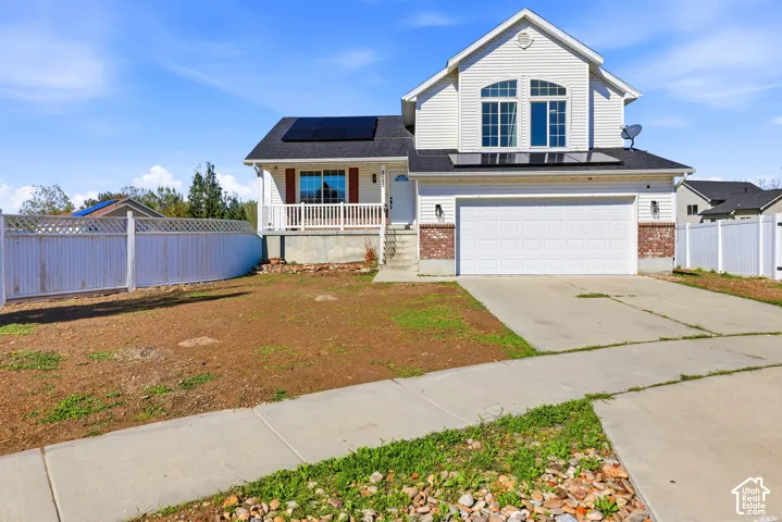 Two-story residence featuring a white vinyl exterior, integrated solar panels, a two-car garage with brick accents, and a raised front porch with white railing