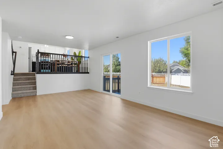 Open-concept living space featuring wood-finish flooring, sliding glass patio doors, neutral wall palette, carpeted staircase with dark railing, and flush-mount ceiling lighting