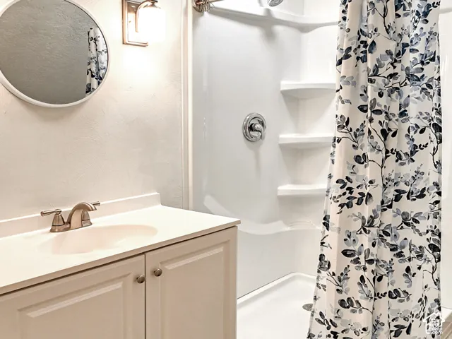 Bathroom featuring a white vanity cabinet with a solid surface countertop, an integrated sink, a brushed nickel faucet, a wall-mounted circular mirror, and a walk-in shower surround with built-in shelving