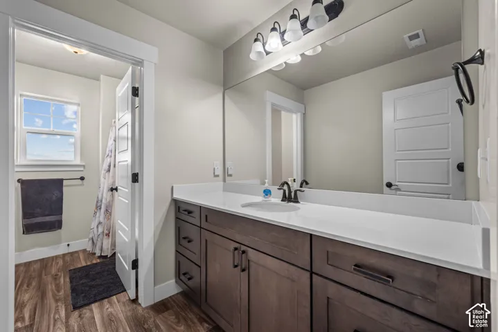 Bathroom vanity featuring stained wood cabinetry, white stone-finish countertop with an integrated basin, matte black hardware, and an expansive wall-to-wall mirror with multi-bulb overhead lighting