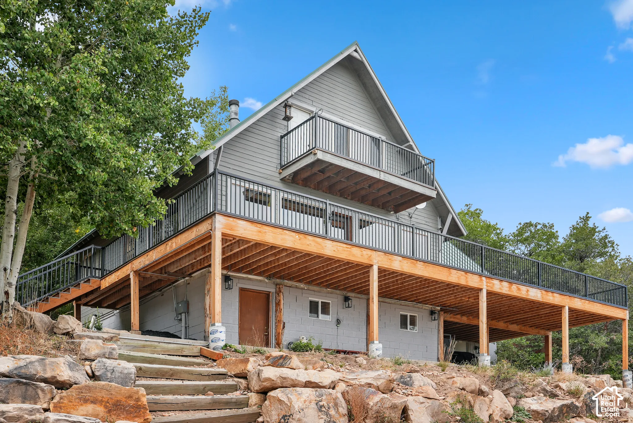 A-frame exterior featuring grey siding, an expansive wrap-around wooden deck, a second-story balcony, black metal railings, and stone masonry hardscaping