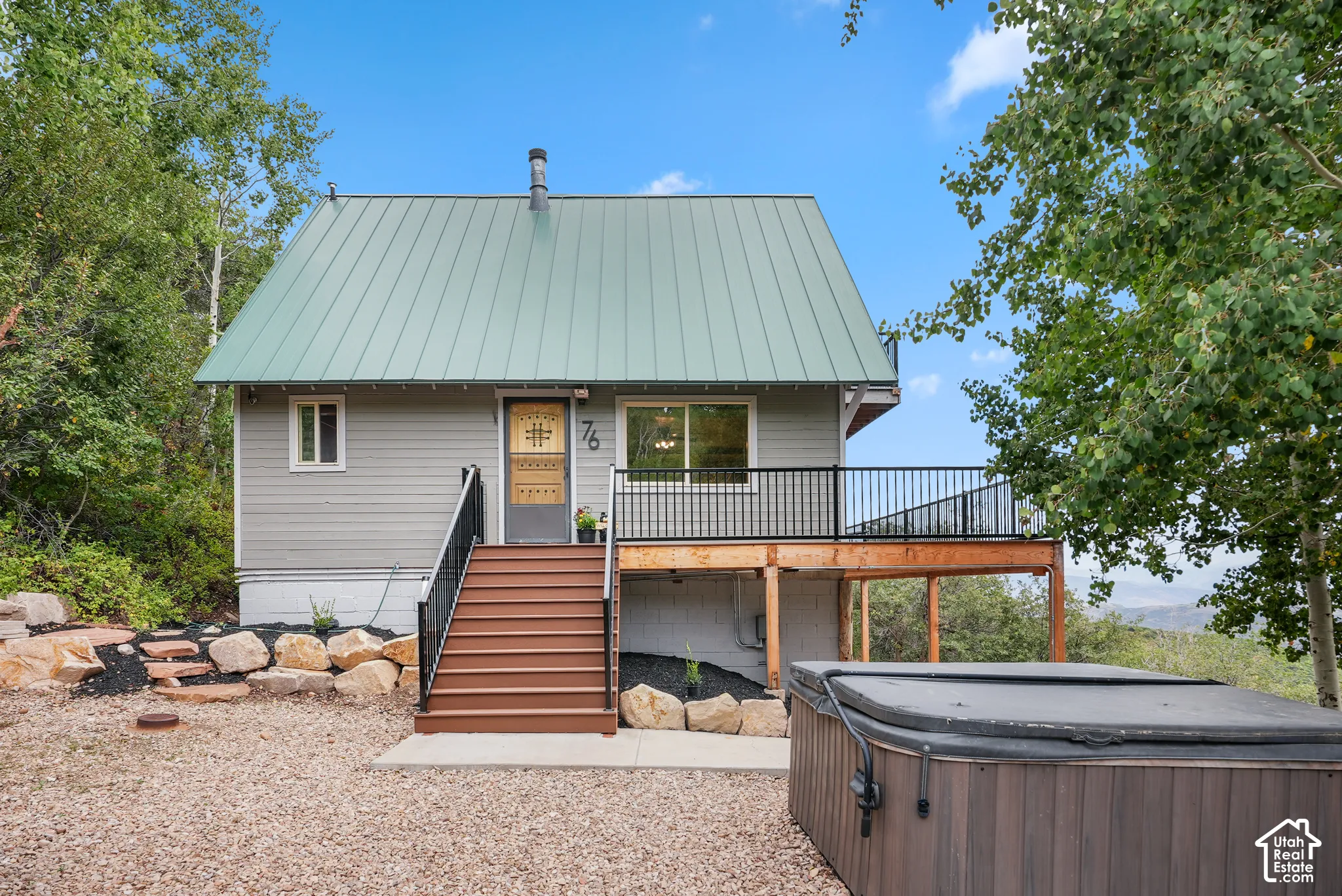 Exterior featuring a green metal gable roof, horizontal siding, raised wooden deck with black metal railing, and rock-lined landscaping