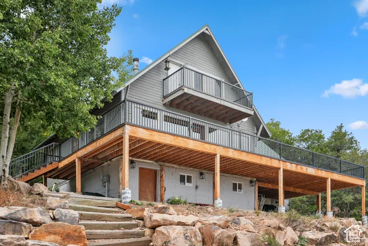 A-frame exterior featuring grey siding, an expansive wrap-around wooden deck, a second-story balcony, black metal railings, and stone masonry hardscaping