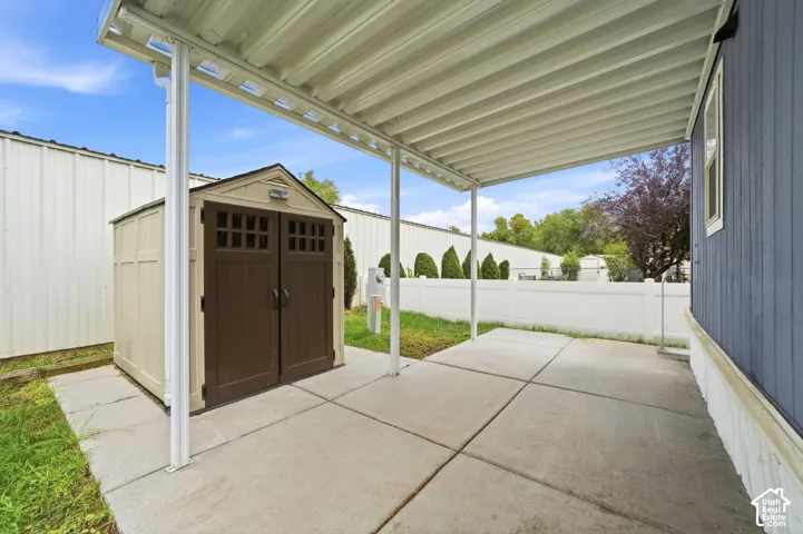 Covered concrete patio with a detached storage structure, enclosed by a white vinyl privacy fence