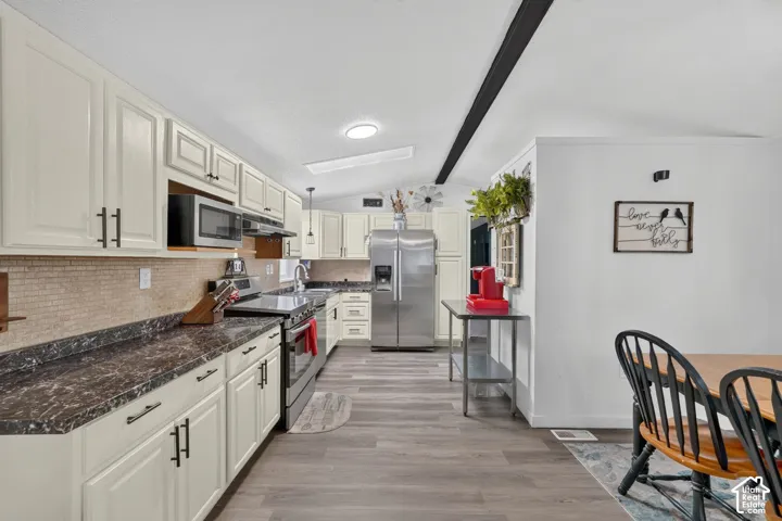 Kitchen featuring off-white cabinetry, dark stone-finish countertops, tiled backsplash, stainless steel appliances, and wood-finish flooring