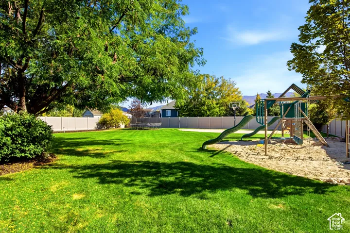 Expansive backyard featuring a manicured lawn, perimeter vinyl fencing, mature shade trees, a built-in play structure, and a sand area