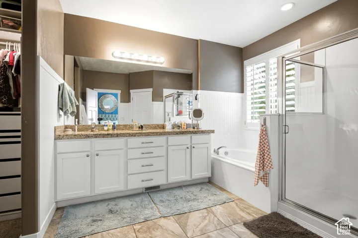Bathroom featuring a double-sink vanity with stone-surface countertop, white shaker-style cabinetry, a soaking tub with beadboard wainscoting, a separate glass-enclosed shower, and large-format tile flooring