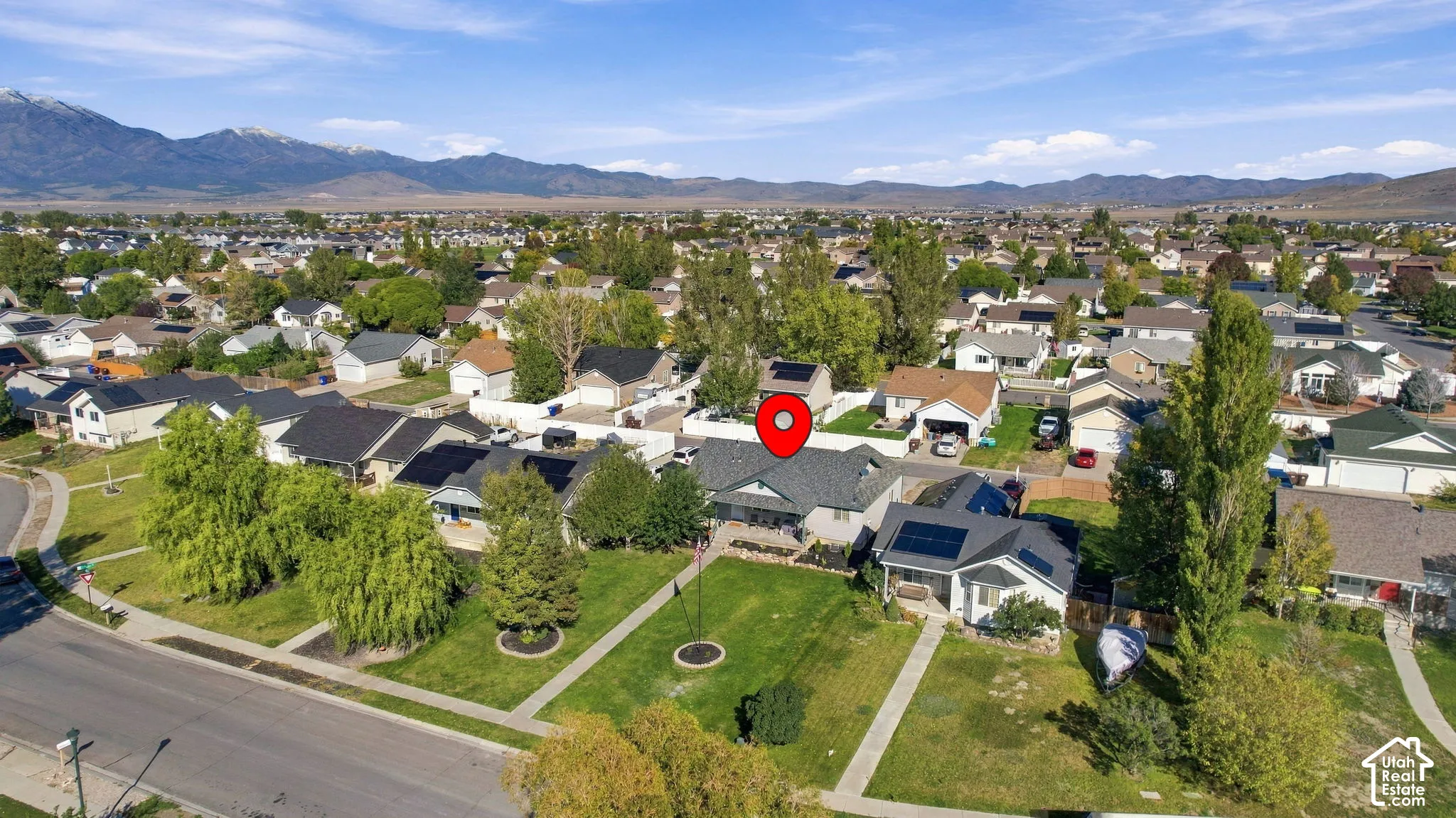 Aerial perspective of a single-family residence featuring a shingled roof, roof-mounted solar panels, an attached garage, and a grass lawn, set against a mountain backdrop