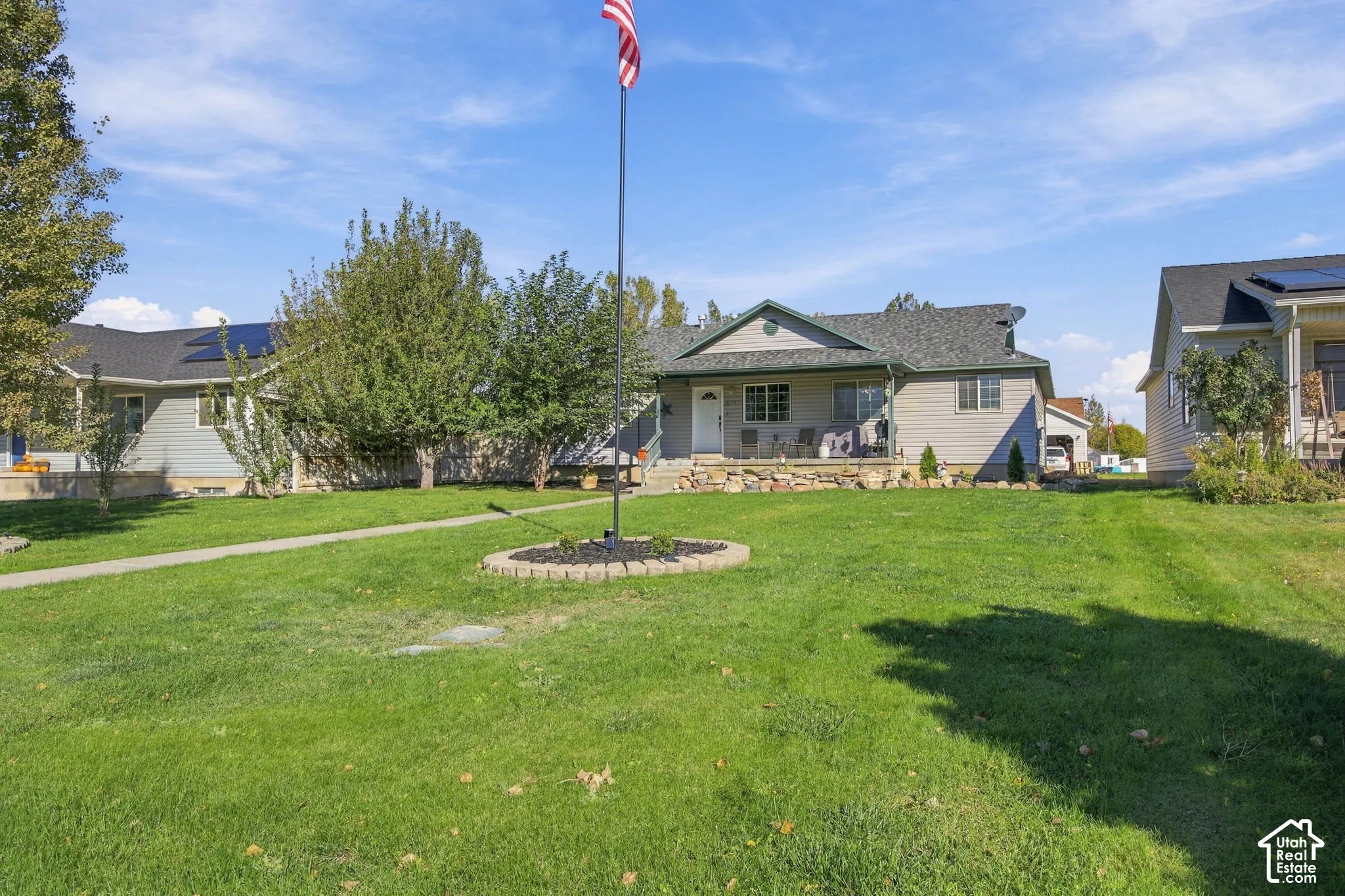 Rambler-style residence featuring horizontal siding, a covered front porch with natural stone masonry, an asphalt shingle roof, solar panels, and a landscaped front lawn with a circular stone-bordered planter