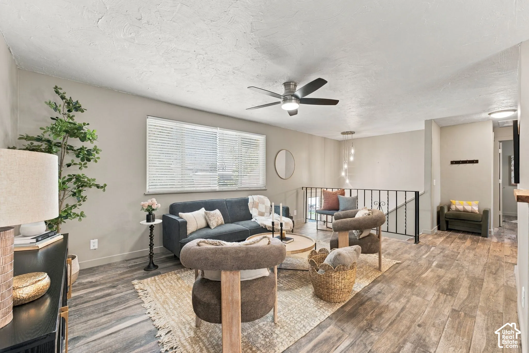 Living space featuring wood-finish flooring, textured ceiling, ceiling fan with integrated lighting, horizontal window blinds, and decorative metal stair railing