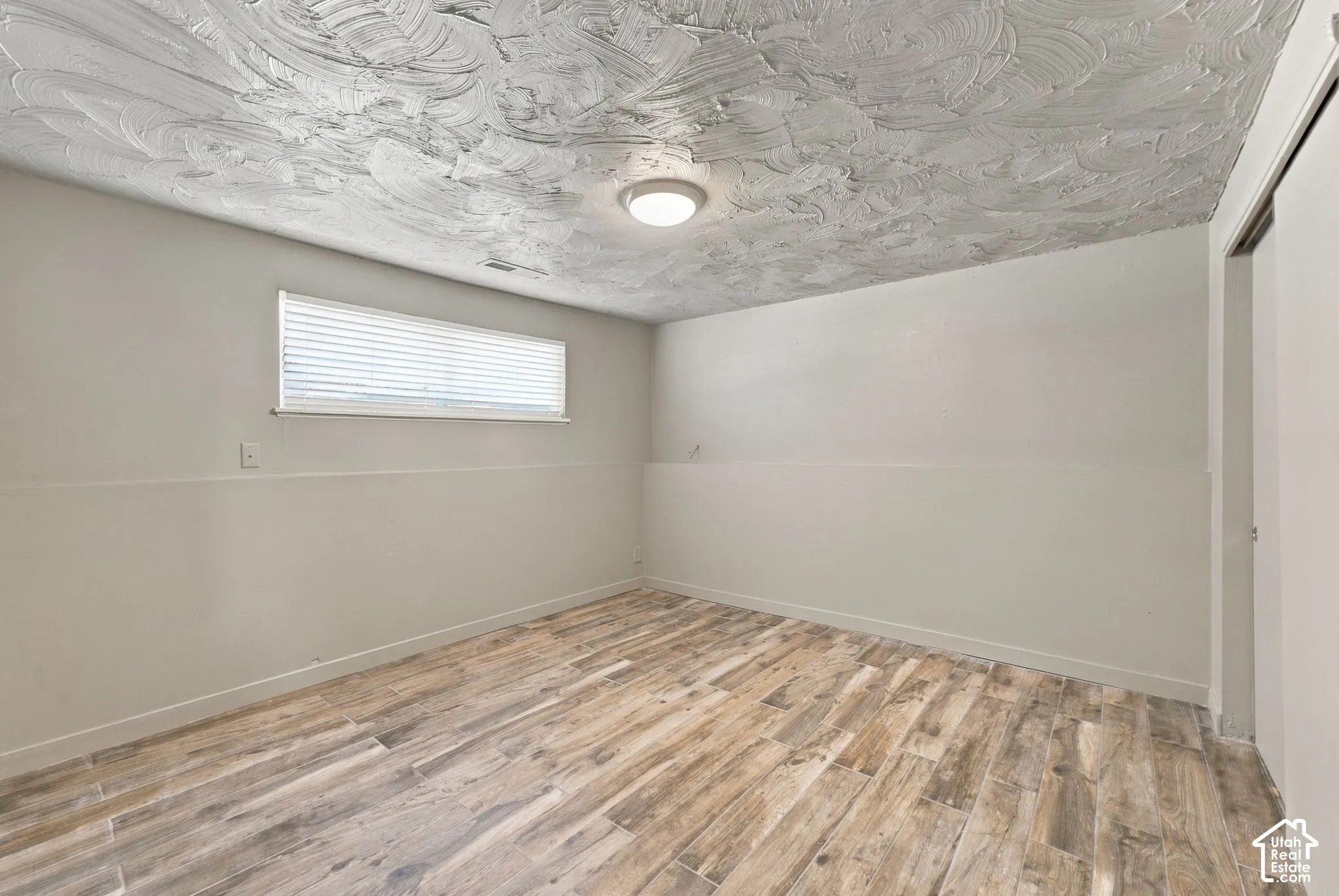 Wood-finish tile flooring, neutral wall paint, ceiling with textured finish, flush-mount lighting, and window with horizontal blinds