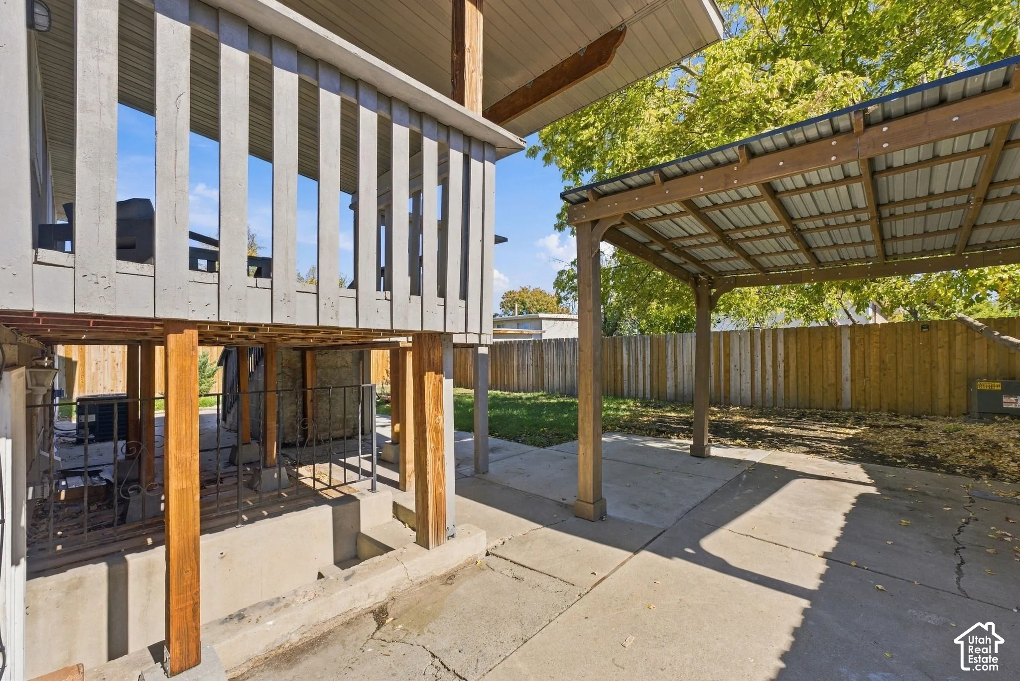Elevated wooden deck with vertical balustrades, timber-framed patio cover with corrugated metal roof, concrete patio, and fenced perimeter