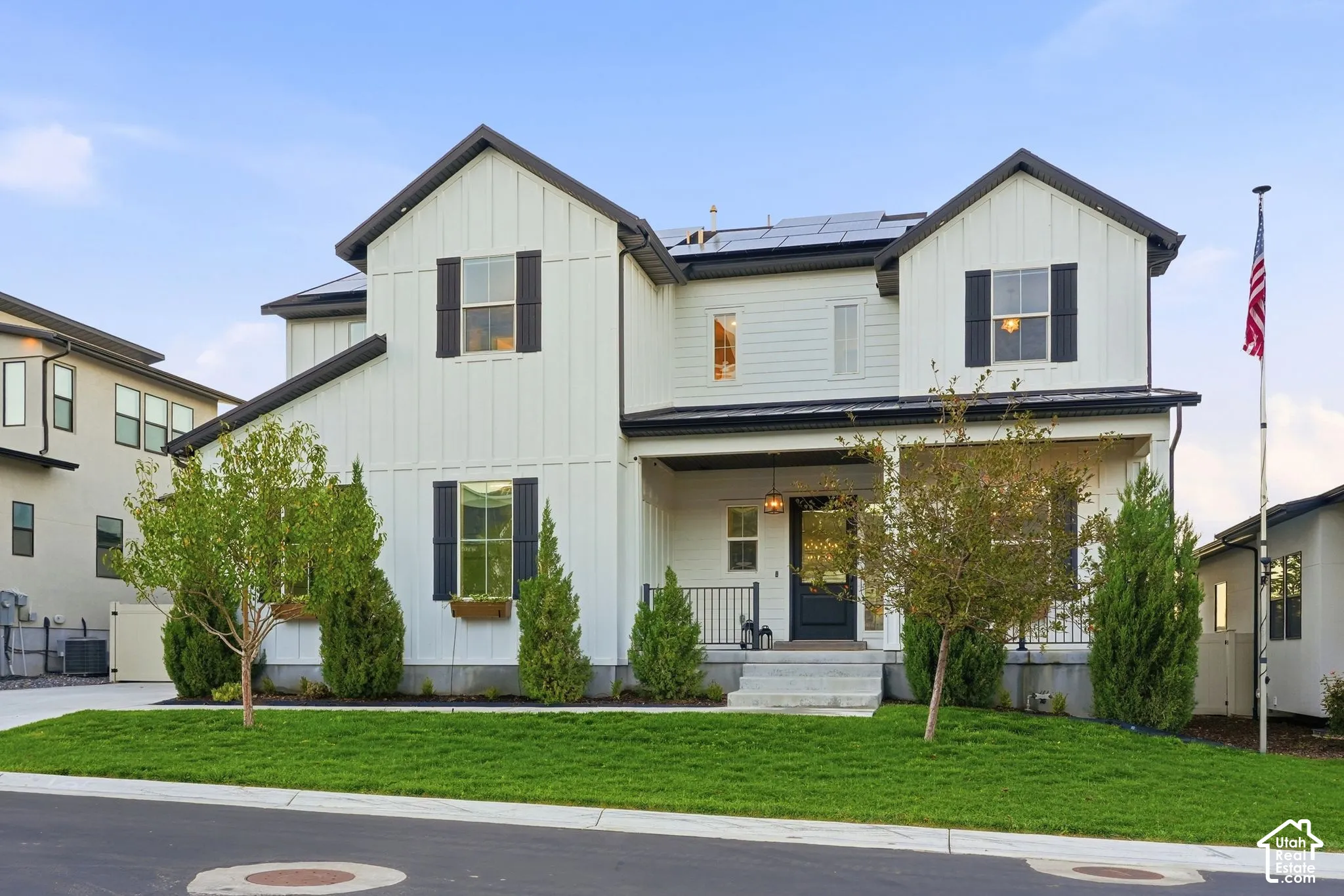 Two-story farmhouse-style exterior featuring vertical board-and-batten siding, dark shutters, a covered front porch with metal roofing, and roof-mounted solar panels