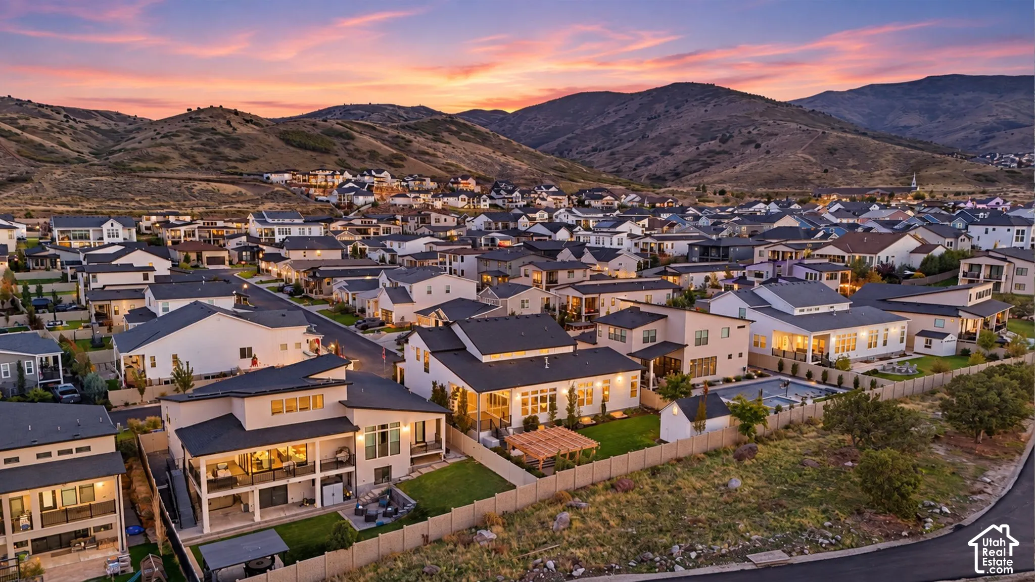Aerial perspective featuring residential lots with manicured lawns, fenced perimeters, covered patios, and pergola structures set against a prominent mountain backdrop