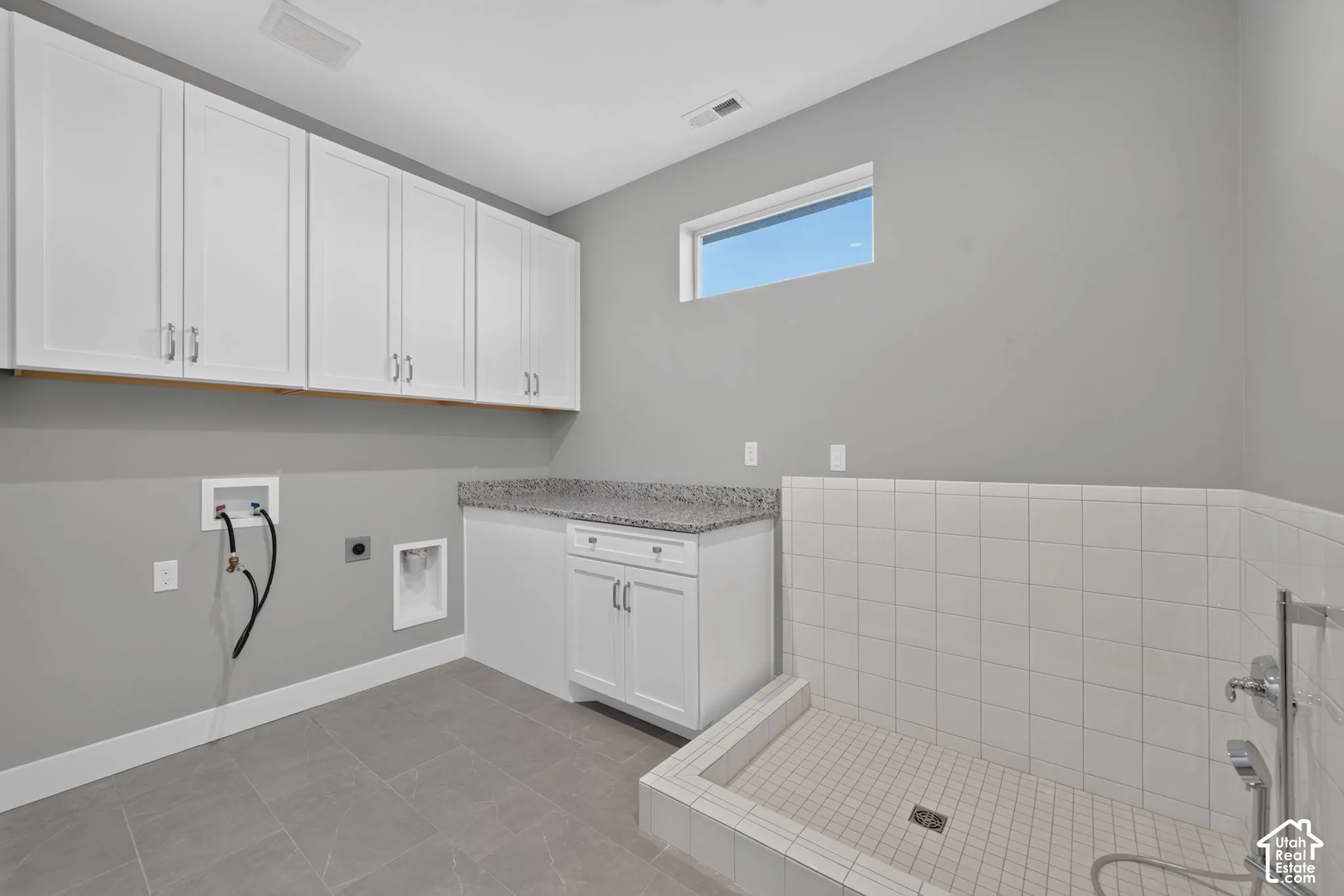 Utility room featuring white shaker-style upper and lower cabinetry, stone-finish countertop, tiled floor, and a walk-in tiled wash station with a chrome faucet