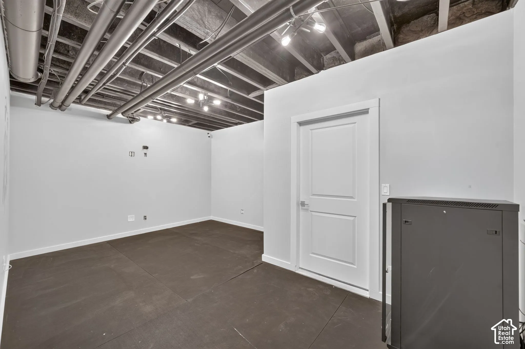 Finished interior space featuring exposed ceiling joists, industrial-style exposed mechanical ductwork, track lighting, dark matte flooring, and a solid-core interior door