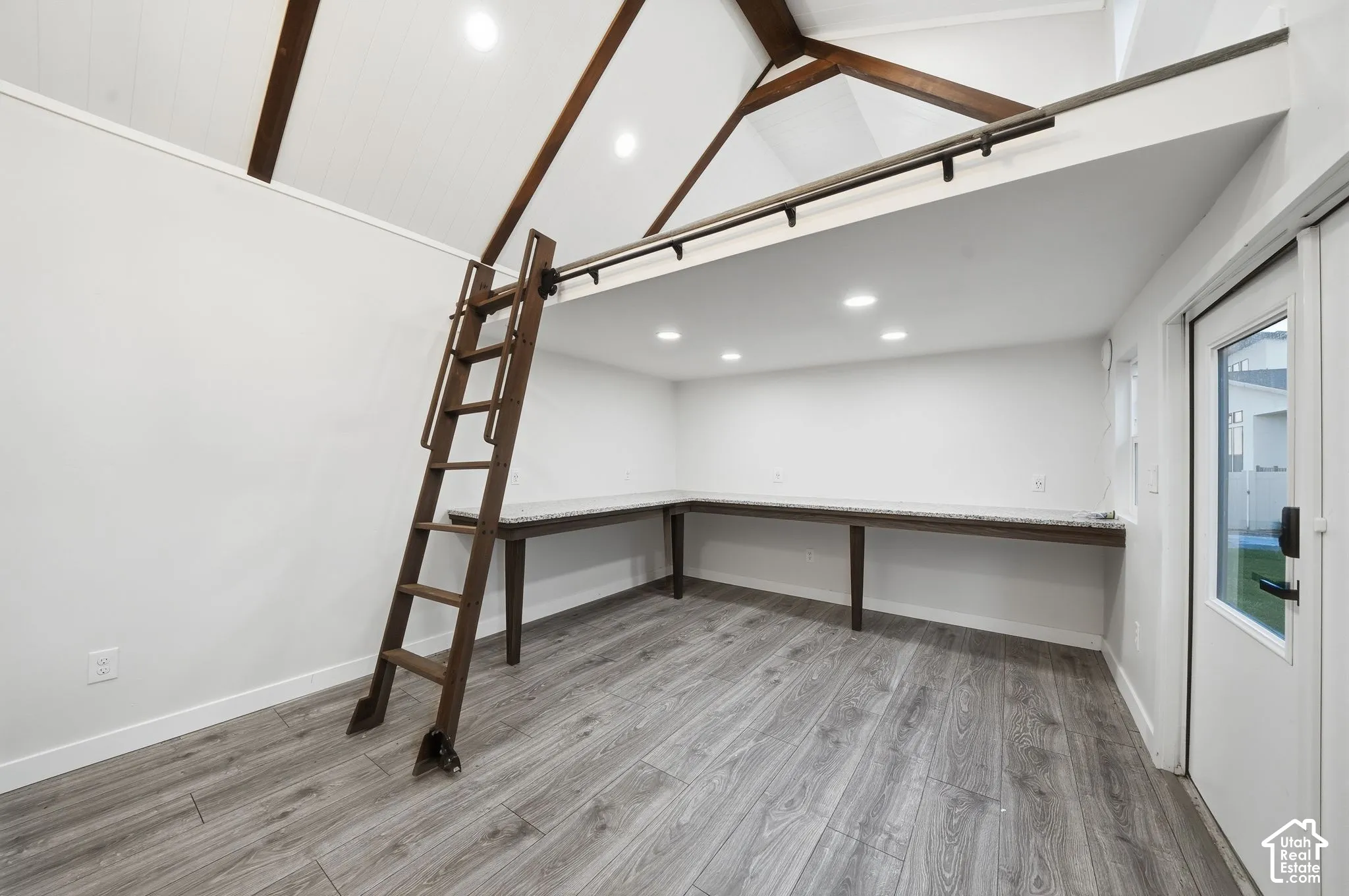 Vaulted ceiling with exposed wood beams, wood-finish flooring, built-in L-shaped desk with stone-finish surface, integrated ladder, and recessed lighting