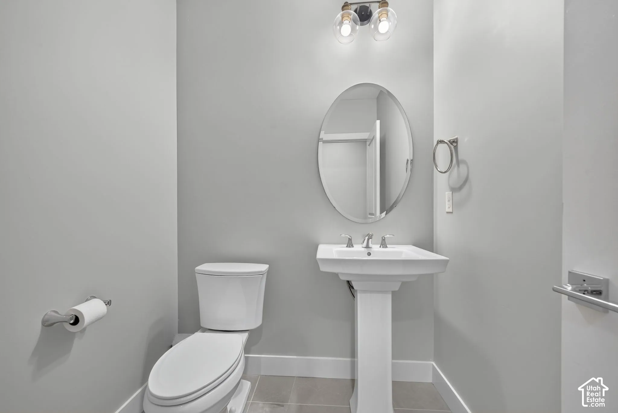 White pedestal sink with chrome faucet, oval wall-mounted mirror, two-light fixture with glass globes, and tile flooring