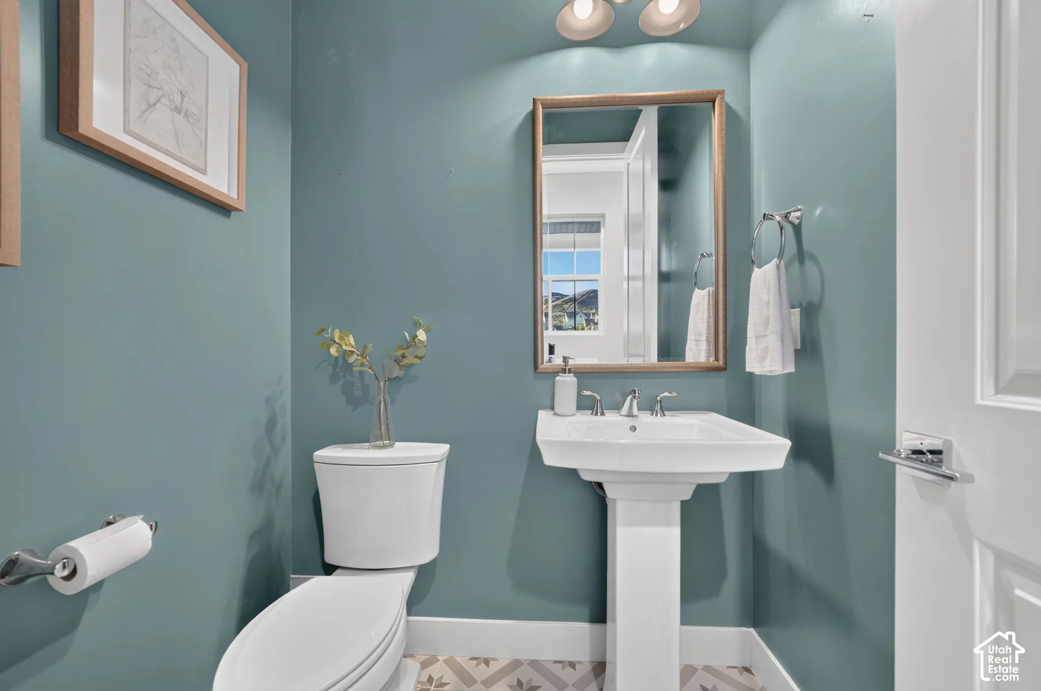 Powder room featuring a pedestal sink, metallic-finished faucet, framed wall mirror, patterned tile flooring, and polished chrome hardware