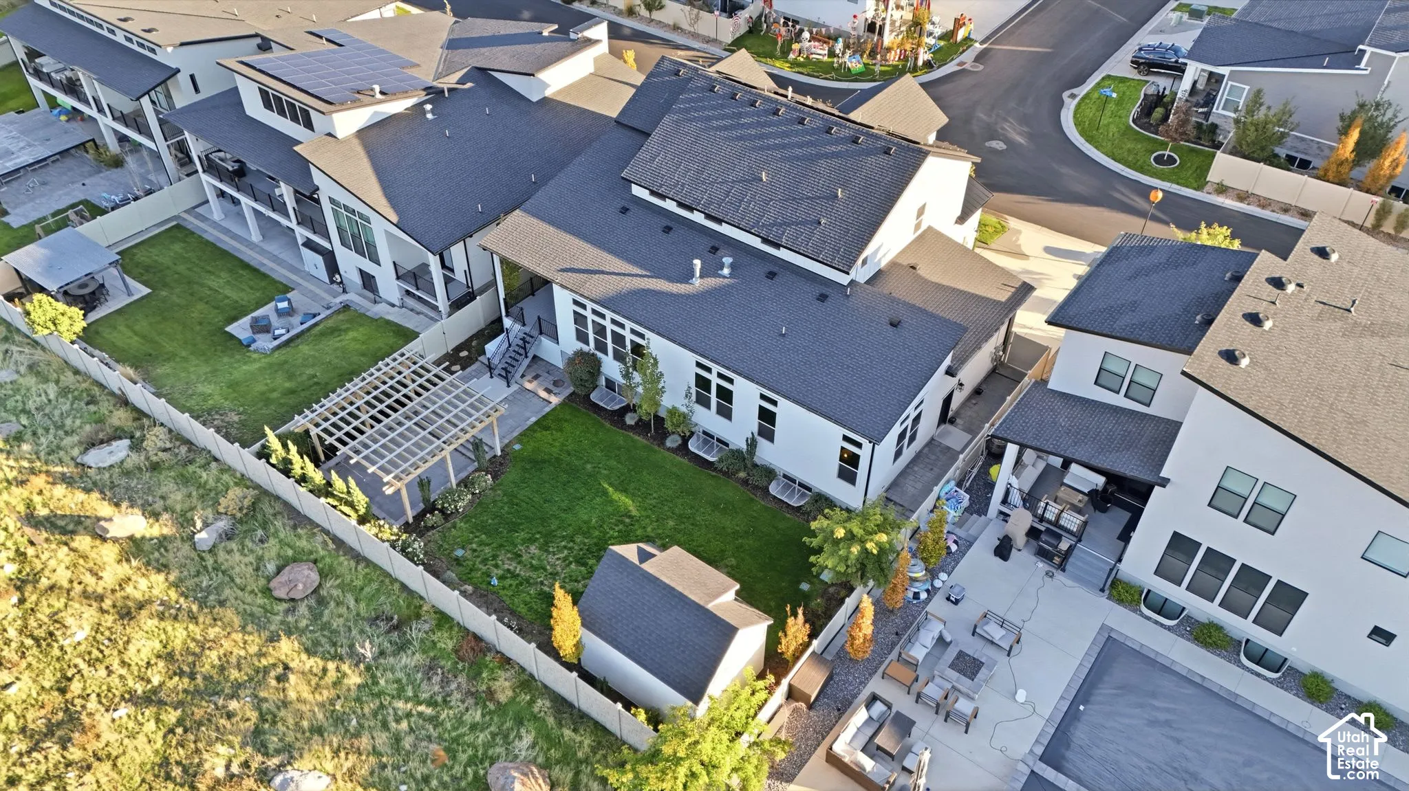 Aerial perspective featuring a multi-story residence with solar panel arrays, a wood-framed pergola over a stone patio, a manicured lawn, and a detached storage structure enclosed by perimeter fencing