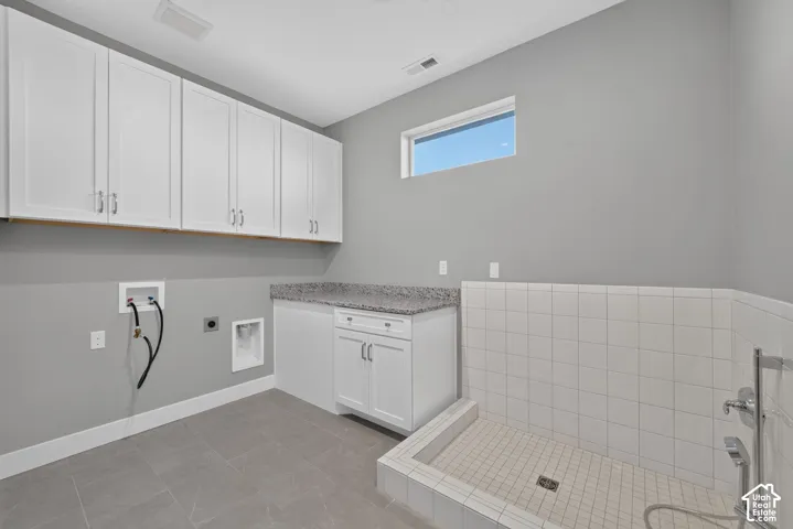 Utility room featuring white shaker-style upper and lower cabinetry, stone-finish countertop, tiled floor, and a walk-in tiled wash station with a chrome faucet