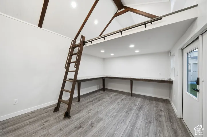 Vaulted ceiling with exposed wood beams, wood-finish flooring, built-in L-shaped desk with stone-finish surface, integrated ladder, and recessed lighting