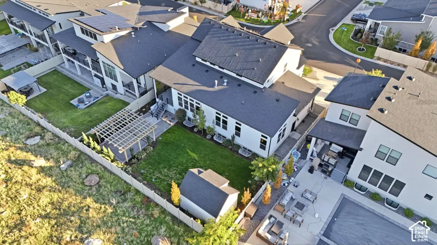 Aerial perspective featuring a multi-story residence with solar panel arrays, a wood-framed pergola over a stone patio, a manicured lawn, and a detached storage structure enclosed by perimeter fencing