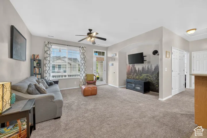 Living area featuring neutral carpeting, a central ceiling fan with integrated lighting, a glass-panel exterior door, and an arched recessed wall niche