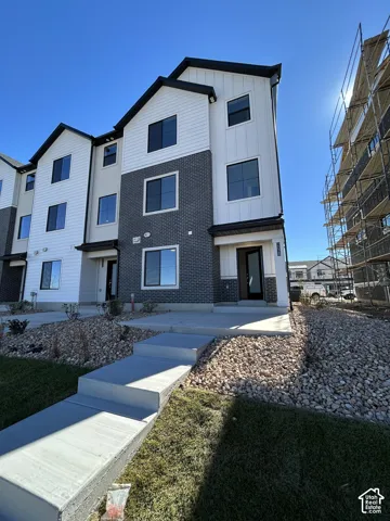 Three-story exterior featuring a mix of horizontal siding and dark brick masonry, vertical gable rooflines, multiple rectangular windows with black frames, and a concrete entrance path